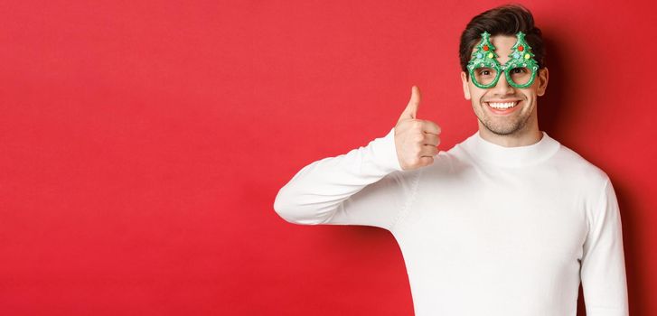 Concept of christmas, winter holidays and celebration. Close-up of handsome smiling man, wearing party glasses and white sweater, showing thumbs-up, recommending new year promo.