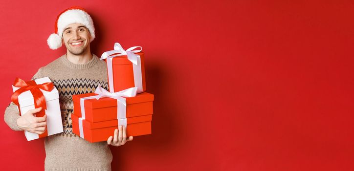 Concept of winter holidays, new year and celebration. Portrait of lovely smiling man prepared gifts for christmas, holding presents and looking at camera heartwarming, red background.