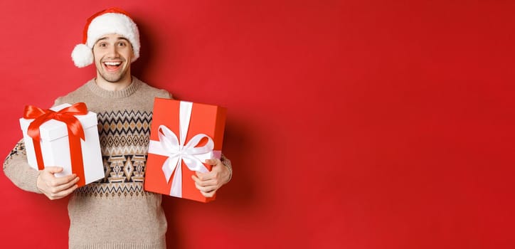 Image of cheerful attractive guy holding christmas presents, standing in santa hat and winter sweater, smiling happy, standing over red background.