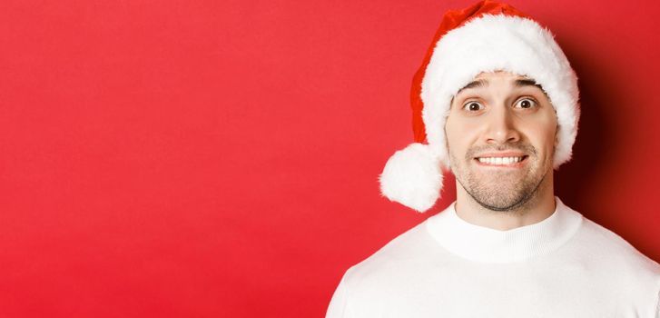 Close-up of handsome guy in santa hat, biting lip and looking with temptation at something he wants, standing over red background.