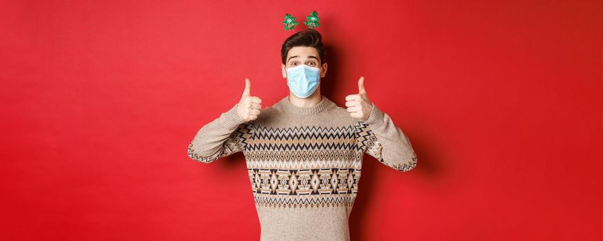 Concept of new year, covid-19 and social distancing. Cheerful caucasian man in sweater and medical mask, celebrating christmas during pandemic, showing thumbs-up, red background.