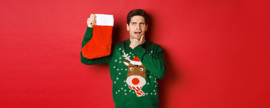 Portrait of thoughtful handsome man in green sweater, holding christmas stocking and thinking, guessing what present is inside, standing over red background.