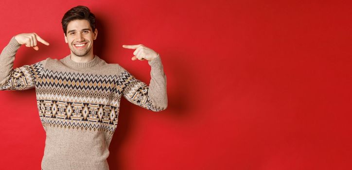 Concept of christmas celebration, winter holidays and lifestyle. Image of happy handsome man in xmas sweater pointing at himself and smiling, being secret santa, standing over red background.