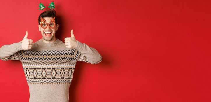 Portrait of satisfied and happy man in christmas sweater and party glasses, showing thumbs-up, wishing happy new year, standing over red background.