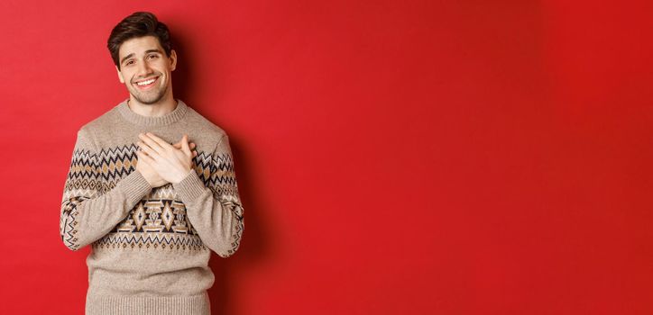 Portrait of touched and happy handsome guy, receiving new year gift, holding hands on heart and smiling, saying thank you, standing in christmas sweater against red background.