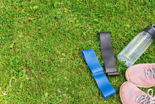 Ladie's fitness rubber bands, water bottle and sneakers on the green grass background, top view. Outdoor training concept