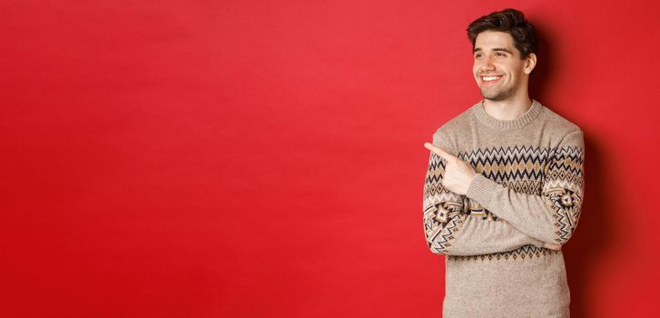 Portrait of attractive caucasian man celebrating new year, winter holidays, pointing finger and looking left with pleased smile, showing christmas advertisement, standing over red background.