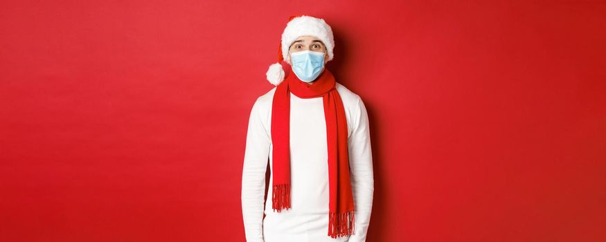 Concept of new year, coronavirus and holidays. Cheerful man celebrating christmas and social distancing, wearing medical mask, santa hat and scarf, standing over red background.