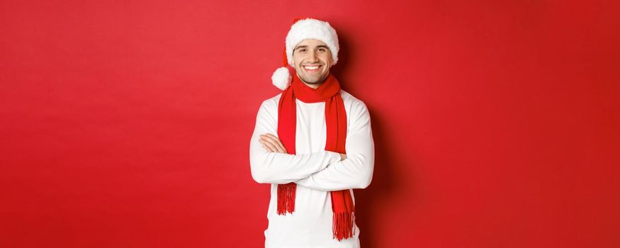 Handsome caucasian guy with bristle, wearing santa hat, scarf and white sweater, cross arms on chest and smiling happy, standing against red background.