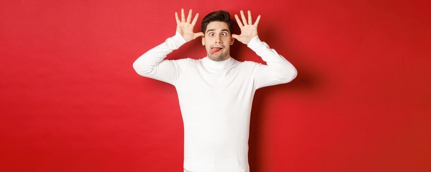 Portrait of funny caucasian guy, showing tongue and making faces, wearing white sweater, standing against red background.