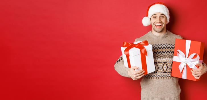 Image of cheerful attractive guy holding christmas presents, standing in santa hat and winter sweater, smiling happy, standing over red background.