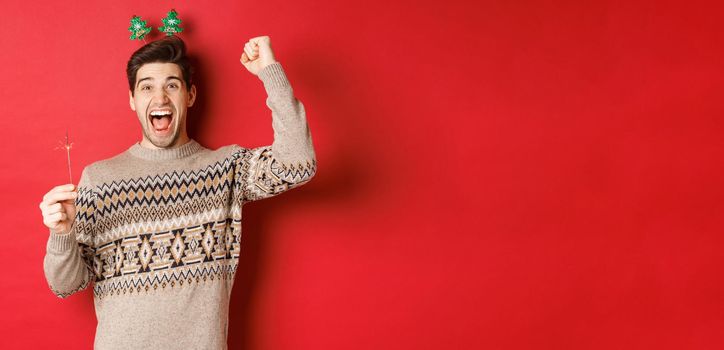 Cheerful guy with a sparkler rejoicing and wishing happy new year, raising hand up and shouting for joy, standing over red background.