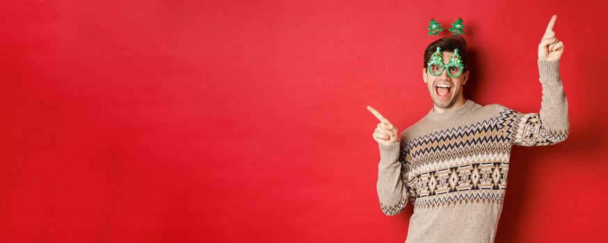 Portrait of joyful handsome guy in party glasses and christmas sweater, dancing and pointing fingers sideways, enjoying new year celebration, standing over red background.