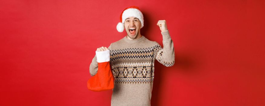 Concept of winter holidays, new year and celebration. Amazed and happy man shouting for joy, found gift inside christmas stocking and cheering, raising hand up and smiling.