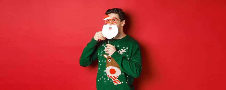 Portrait of surprised man in green christmas sweater, holding funny santa claus mask, celebrating new year, looking left amazed, sanding over red background.