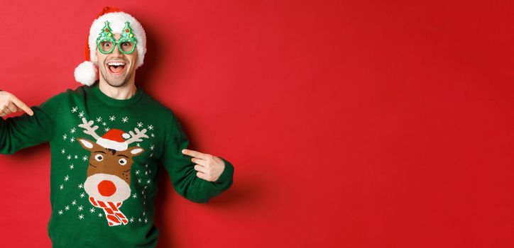 Image of happy man in party glasses and santa hat, pointing at his christmas sweater and smiling, standing over red background.