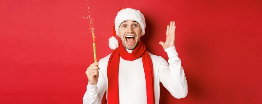 Concept of christmas, winter holidays and celebration. Handsome man celebrating new year and having fun, holding sparkler and smiling, wearing santa hat, standing over red background.