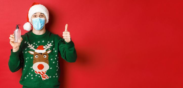 Concept of new year, coronavirus and social distancing. Portrait of cheerful man in santa hat, christmas sweater and medical mask, showing thumb-up and recommending hand sanitizer.