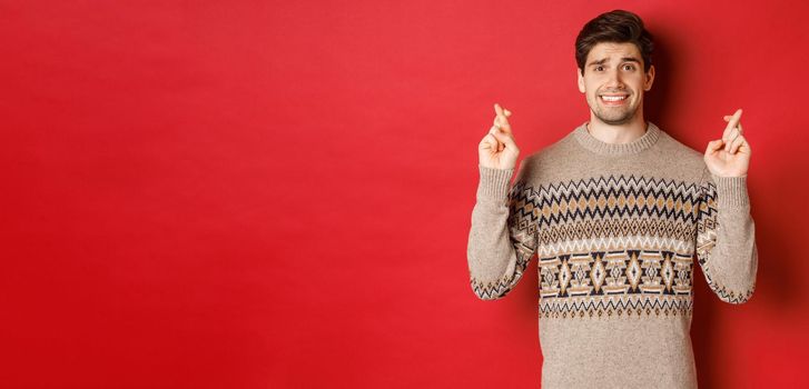 Image of hopeful and worried man in christmas sweater waiting for something, cross fingers for good luck and making wish, nervous about new year gift, red background.