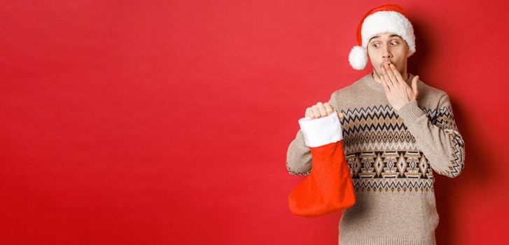 Concept of winter holidays, new year and celebration. Surprised adult man holding christmas stocking with presents inside, looking amazed at bag, standing over red background in santa hat.