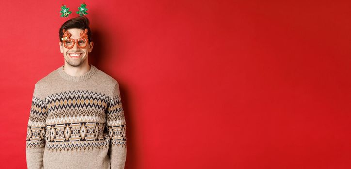 Portrait of handsome bearded guy in party glasses and winter sweater, smiling pleased, celebrating new year, wishing merry christmas, standing over red background.