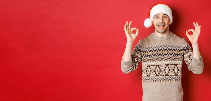 Portrait of happy and pleased handsome man, wearing winter sweater and santa hat, showing okay signs, praise good christmas party, standing over red background.