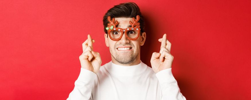 Concept of winter holidays, christmas and celebration. Close-up of nervous man in party glasses, crossing fingers for good luck and pleading, making a wish, standing over red background.