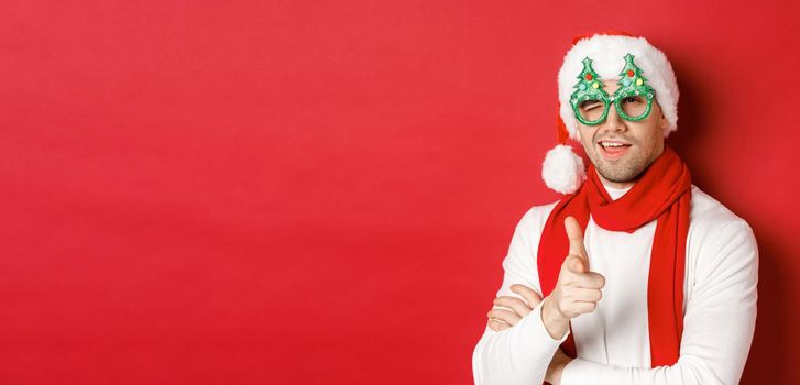 Concept of christmas, winter holidays and celebration. Close-up of cheeky young man in santa hat and party glasses, smiling and pointing finger gun at camera, standing over red background.