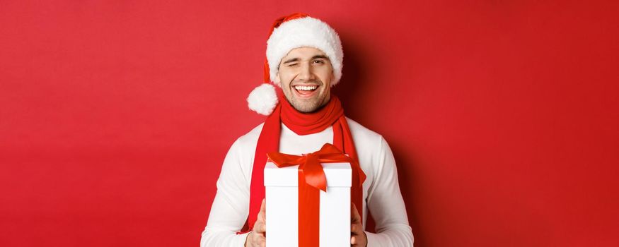 Concept of winter holidays, christmas and lifestyle. Handsome cheeky man in santa hat and scarf, holding present and smiling, winking at camera, standing over red background.