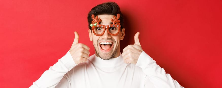 Concept of winter holidays, christmas and celebration. Close-up of excited handsome man in party glasses, smiling and showing thumbs-up in approval, standing over red background.