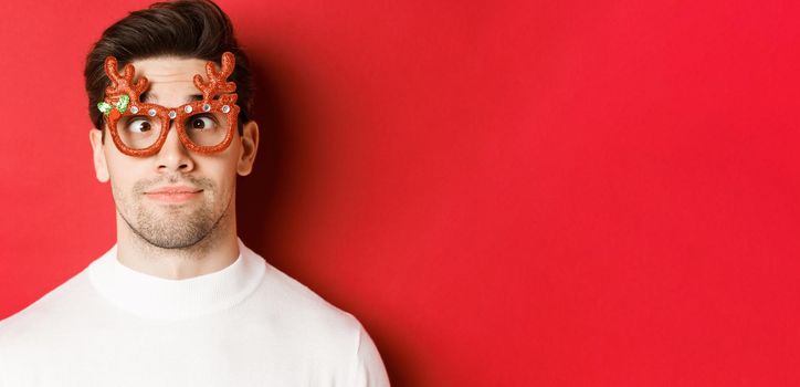 Concept of winter holidays, christmas and celebration. Close-up of funny brunette in party glasses, squinting and making faces, standing over red background.