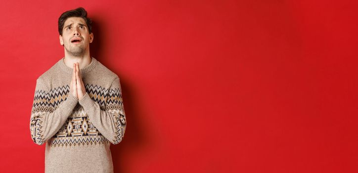 Image of nervous and hopeful man praying to God, begging for help with christmas, wearing winter sweater, looking up and pleading, standing over red background.