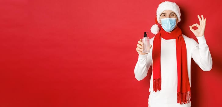 Concept of covid-19, christmas and holidays during pandemic. Attractive man in santa hat and medical mask, showing okay sign while recommending hand sanitizer, standing over red background.