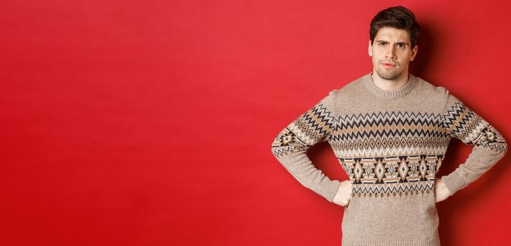 Portrait of doubtful and disappointed man, wearing christmas sweater, frowning and looking displeased at you, standing against red background.