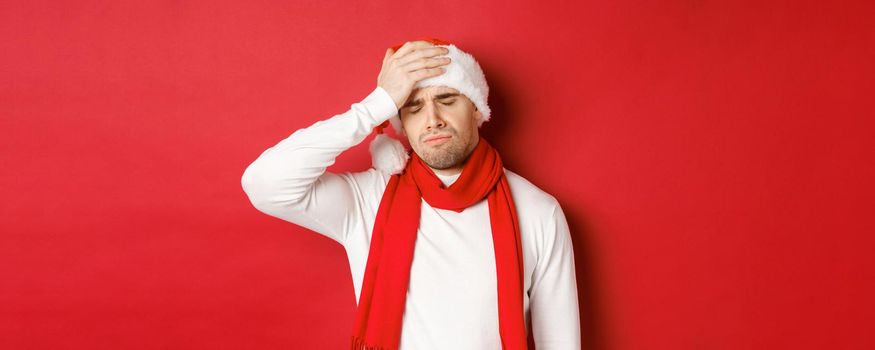 Concept of christmas, winter holidays and celebration. Portrait of sad and frustrated man in santa hat, feeling bad, standing over red background.