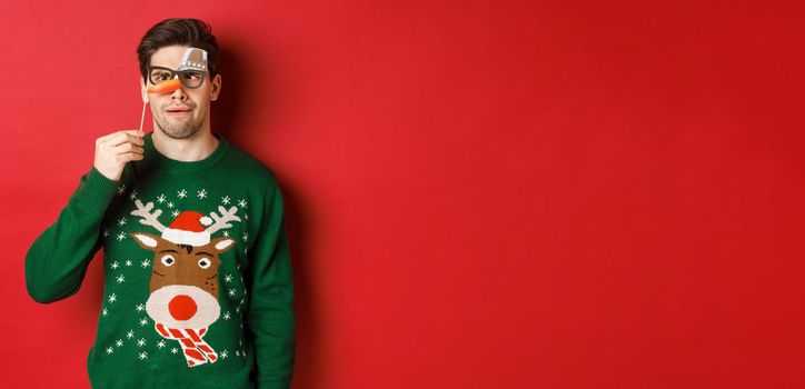 Funny man in christmas sweater and party mask, celebrating winter holidays, showing funny faces, standing over red background.