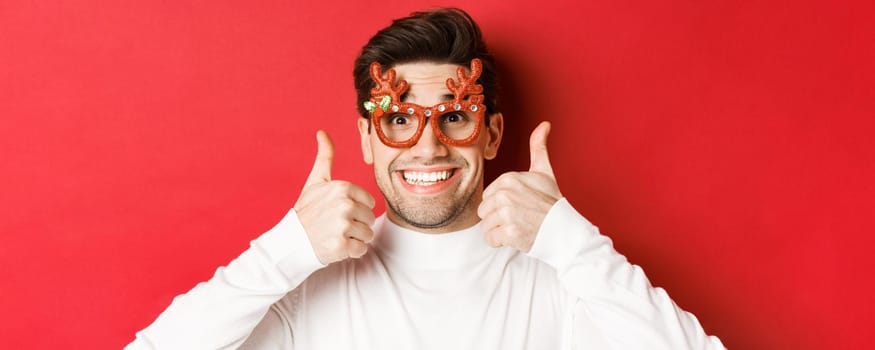 Concept of winter holidays, christmas and celebration. Close-up of excited handsome man in party glasses, smiling and showing thumbs-up in approval, standing over red background.