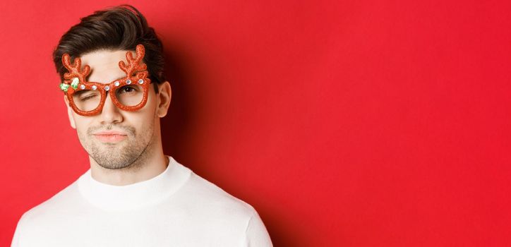 Concept of winter holidays, christmas and celebration. Close-up of cheeky attractive man in party glasses, winking and looking sassy, standing over red background.