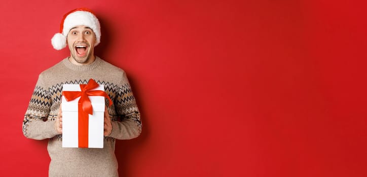 Image of excited handsome man receiving christmas gift, wearing santa hat and winter sweater, shouting for joy, holding present and standing over red background.