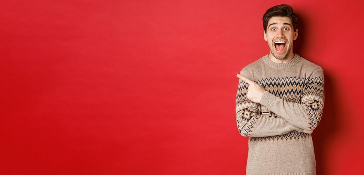 Portrait of amazed handsome guy, wearing christmas sweater, pointing finger at upper left corner logo and looking excited, showing awesome new year promo offer, standing over red background.