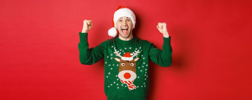 Portrait of cheerful attractive man celebrating new year, wearing green sweater and santa hat, shouting for joy, winning or triumphing, standing over red background.