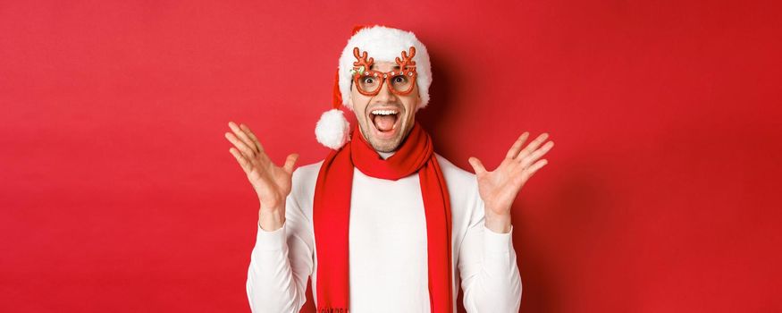 Concept of christmas, winter holidays and celebration. Image of surprised and happy man looking amazed, wearing party glasses and enjoying new year, standing over red background.