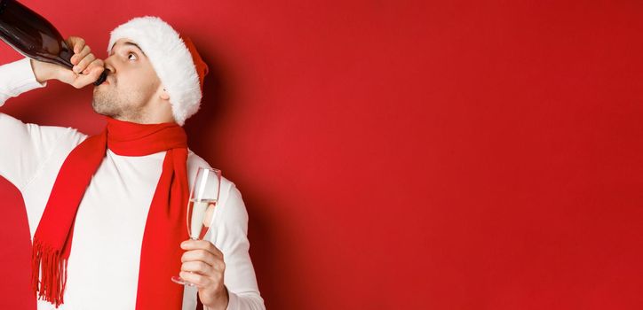 Concept of winter holidays, christmas and lifestyle. Man getting drunk on new year party, drinking champagne from bottle, wearing santa hat, standing over red background.