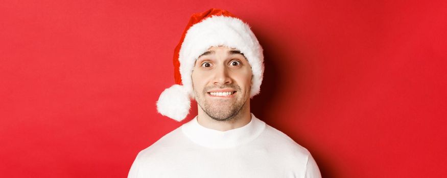 Close-up of handsome guy in santa hat, biting lip and looking with temptation at something he wants, standing over red background.