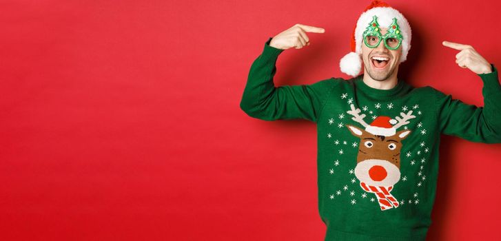 Image of excited smiling guy pointing at party glasses and celebrating new year, standing joyful in green sweater and santa hat against red background.