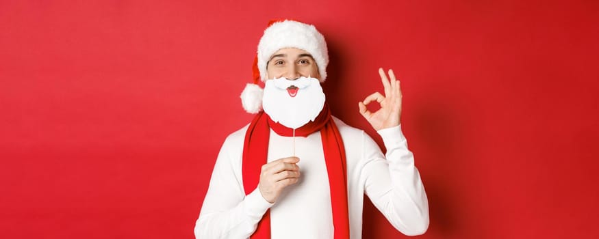 Concept of christmas, winter holidays and celebration. Pleased handsome man in santa hat, holding long white beard mask and showing okay sign, standing over red background.