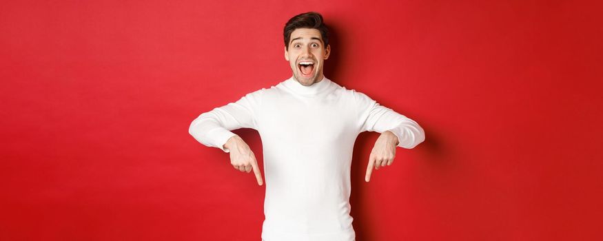 Image of amazed handsome guy in white sweater, reacting excited to christmas advertisement, pointing fingers down, showing logo, standing over red background.