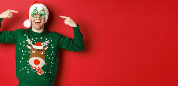 Image of excited smiling guy pointing at party glasses and celebrating new year, standing joyful in green sweater and santa hat against red background.