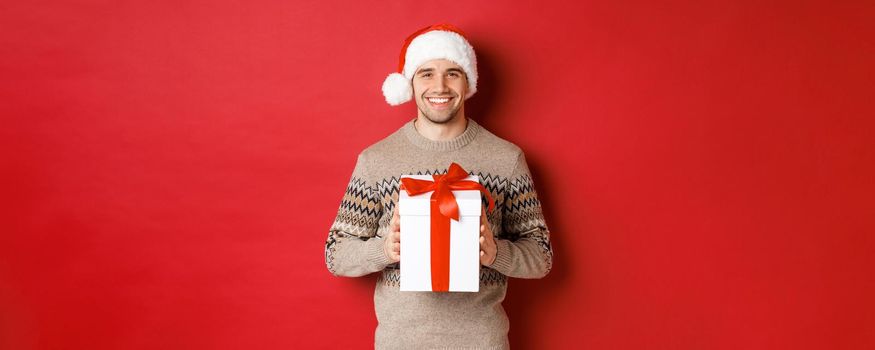 Image of handsome smiling man in santa hat and winter sweater, holding a present, giving christmas gift and wishing happy holidays, standing over red background.