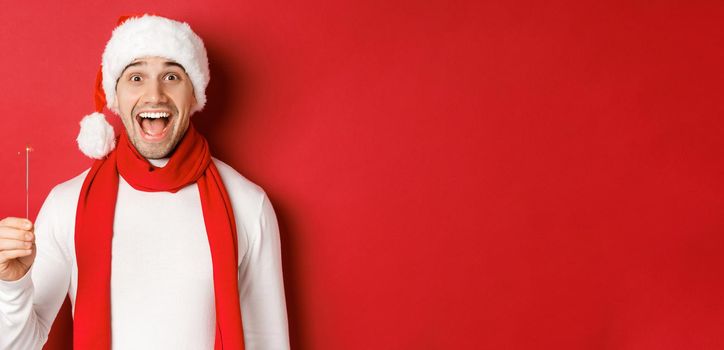 Concept of christmas, winter holidays and celebration. Close-up of happy handsome man in santa hat and scarf, having fun on new year party, holding sparkler and smiling, red background.